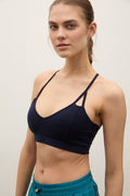 A woman wearing a dark navy blue sports bra with thin crossover straps, paired with teal athletic shorts, against a plain light gray background.