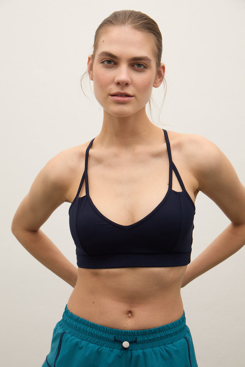 A woman modeling a dark navy blue sports bra with thin straps and side cutouts, paired with teal athletic shorts, against a plain light gray background.