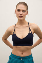 A woman modeling a dark navy blue sports bra with thin straps and side cutouts, paired with teal athletic shorts, against a plain light gray background.