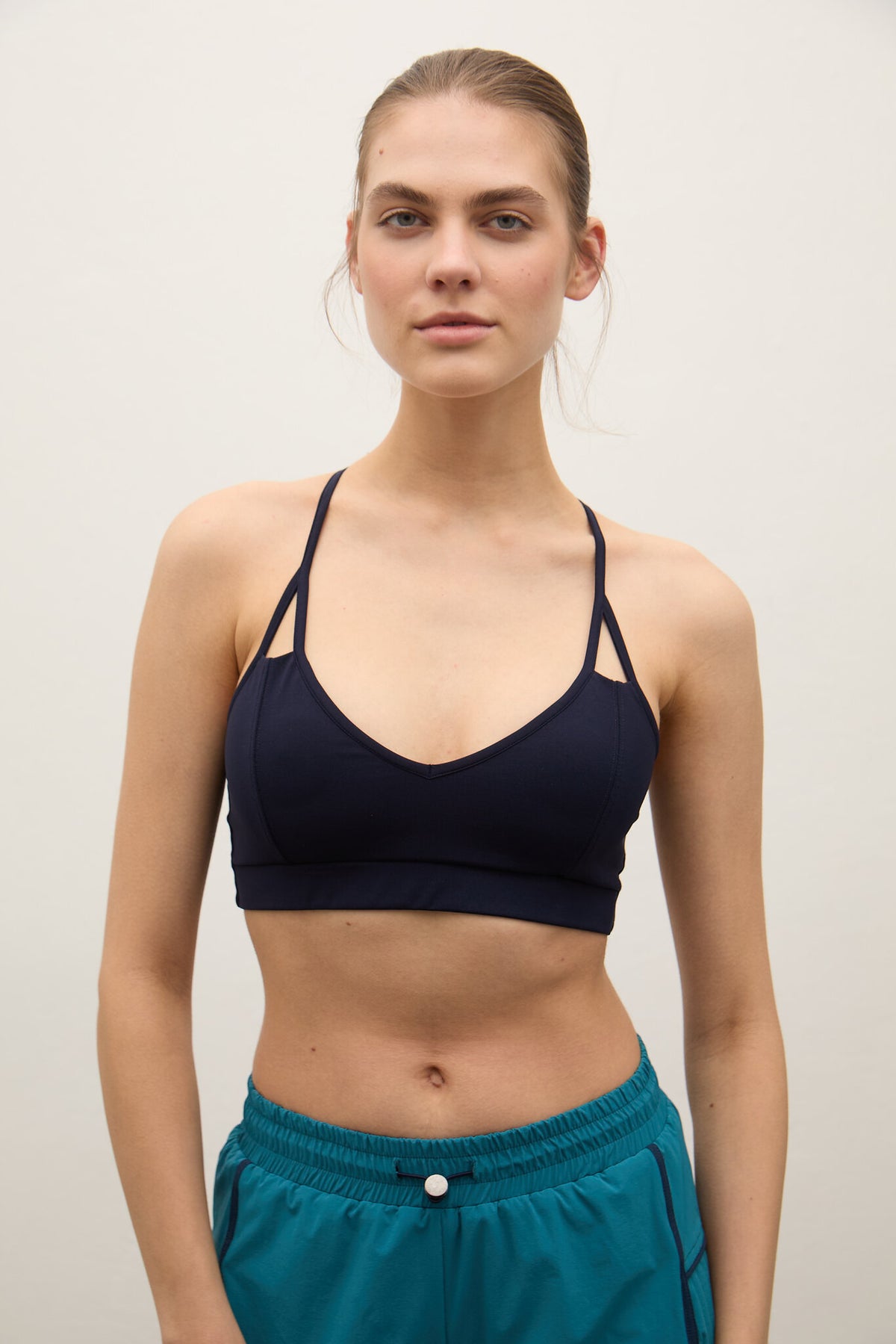 A woman modeling a dark navy blue sports bra with thin straps and teal athletic shorts against a plain light background.