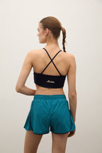 A woman shown from the back wearing a black sports bra with crisscross straps and a small gold logo, paired with teal athletic shorts, against a plain light gray studio background.