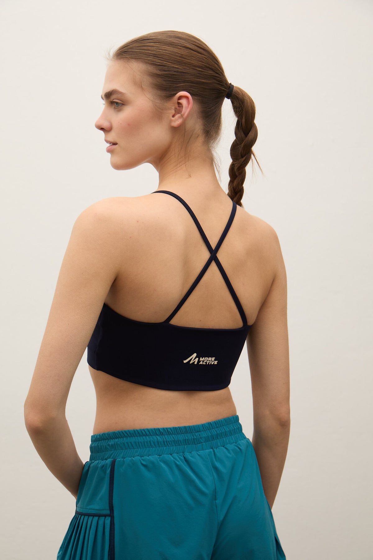 A woman shown from the back wearing a black sports bra with thin crisscross straps and a small gold logo, paired with teal athletic shorts, against a plain light gray studio background.