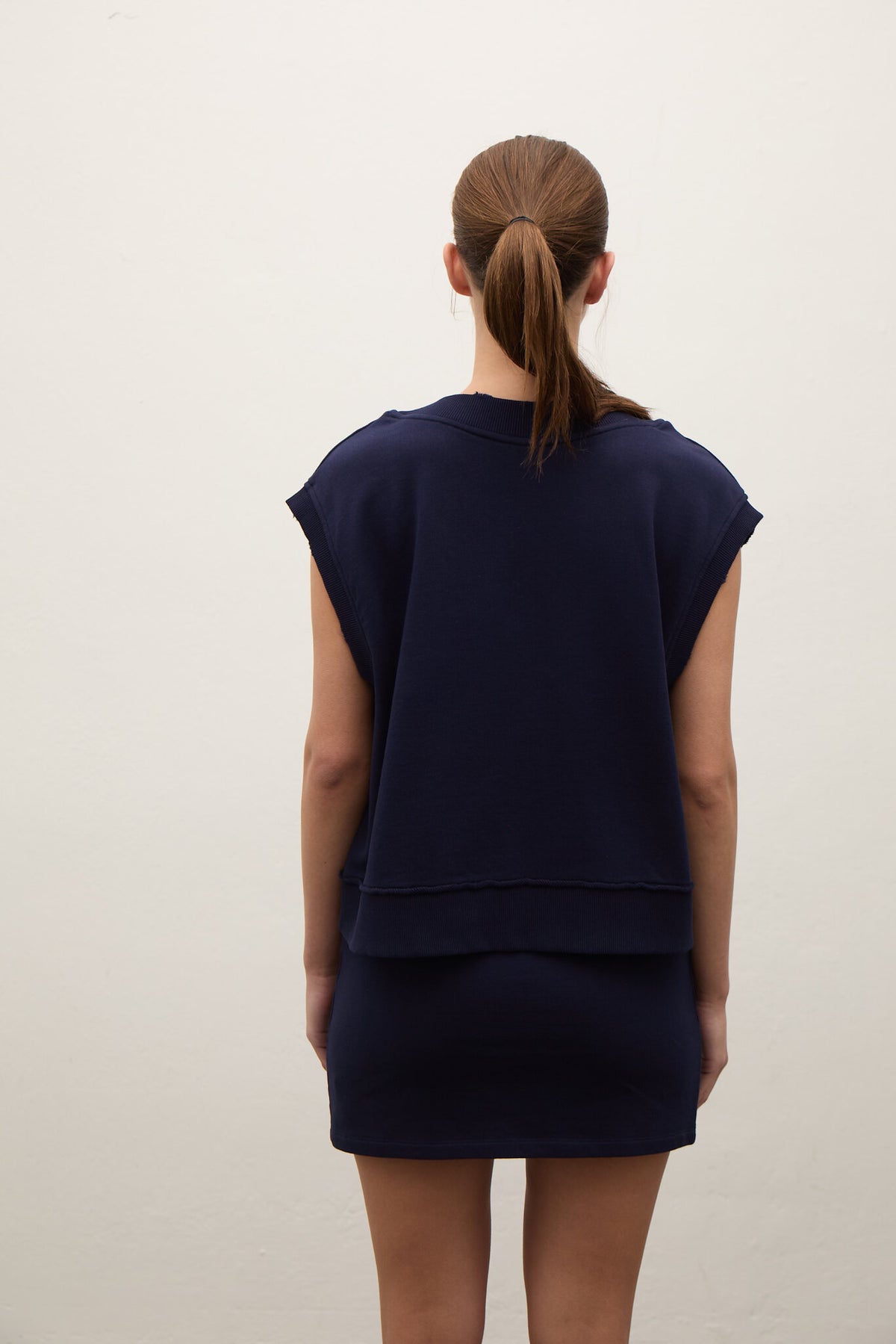 A woman viewed from behind wearing a navy blue sleeveless top and matching mini skirt against a plain off-white wall.