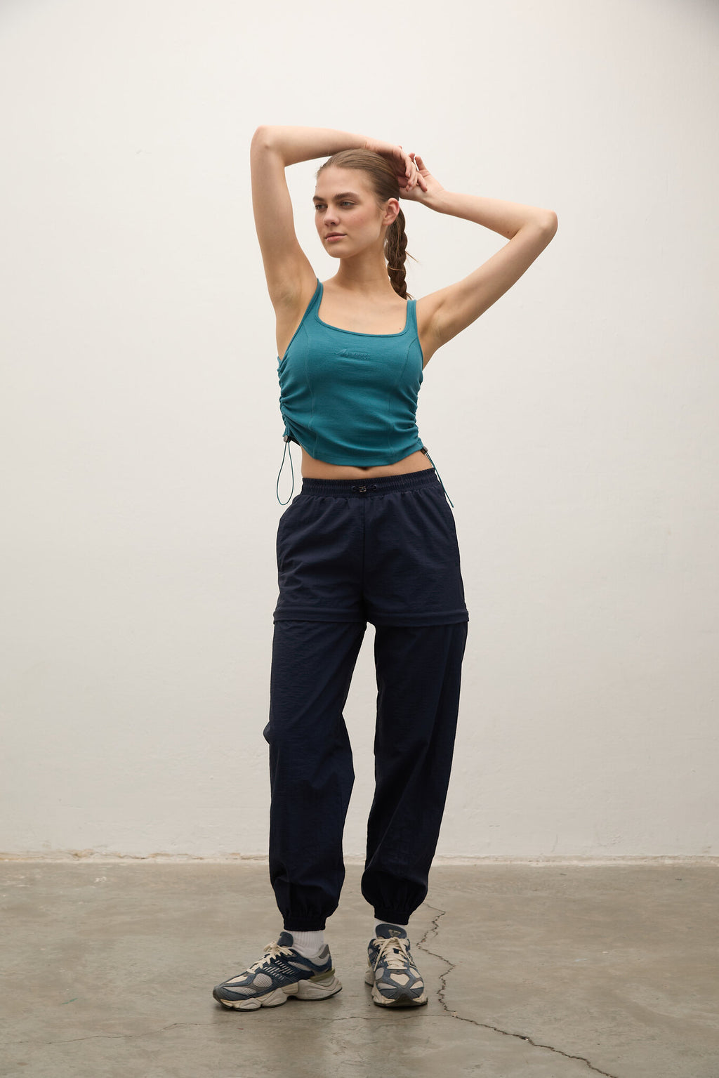 A woman modeling a teal cropped tank top and dark navy jogger sweatpants with gray sneakers, standing against a plain off-white wall.