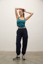 A woman modeling a teal cropped tank top and dark navy jogger sweatpants with gray sneakers, standing against a plain off-white wall.