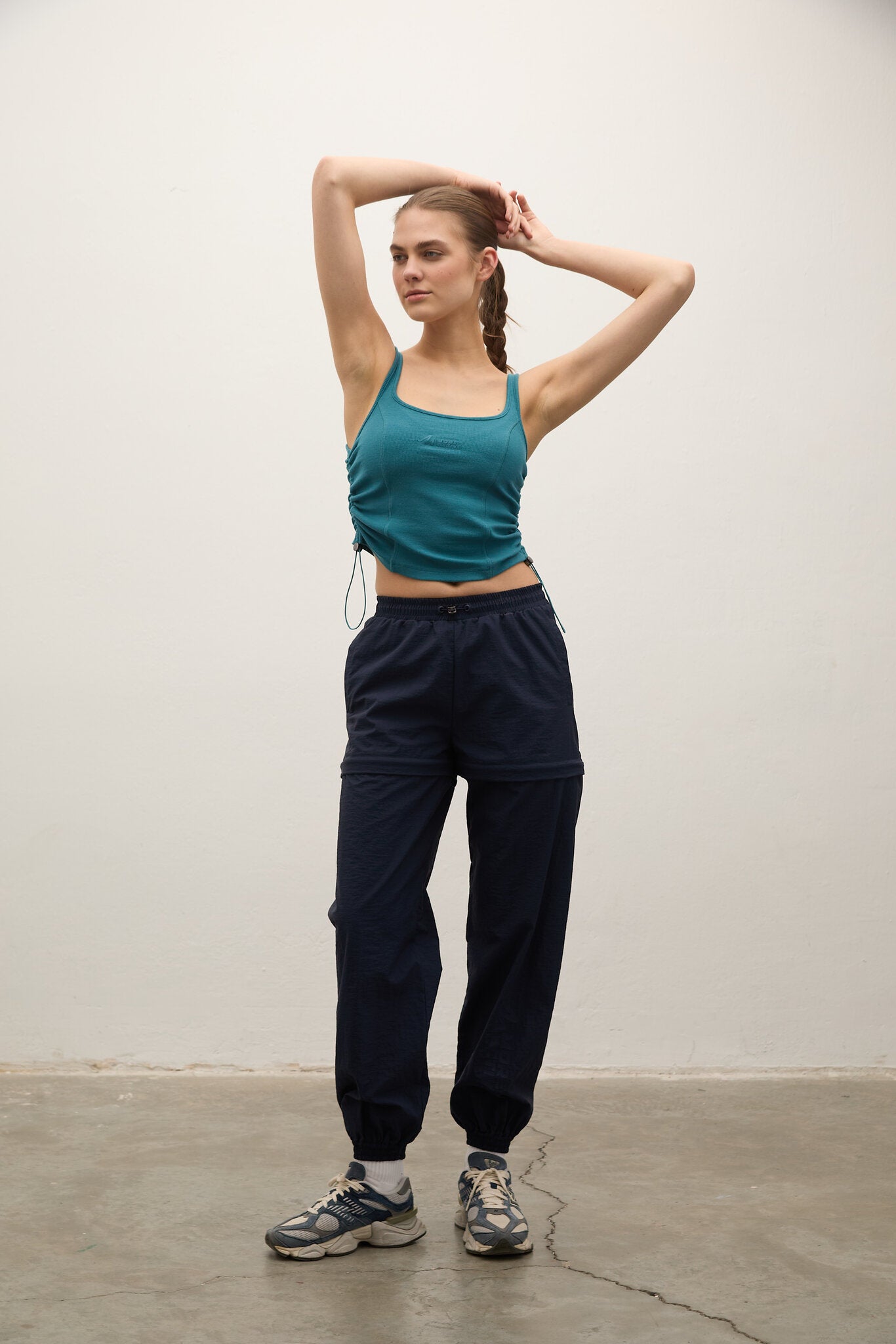 A woman modeling a teal cropped tank top and dark navy jogger sweatpants with gray sneakers, standing against a plain off-white wall.