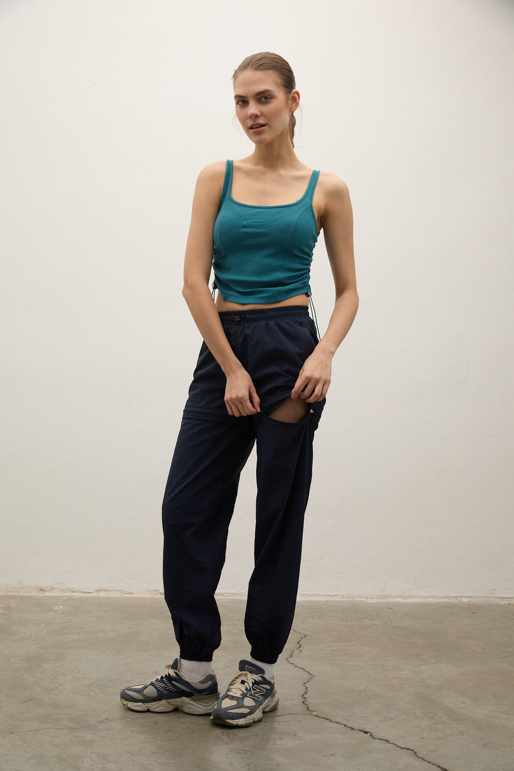 Woman wearing a teal tank top and black pants standing against a white wall.