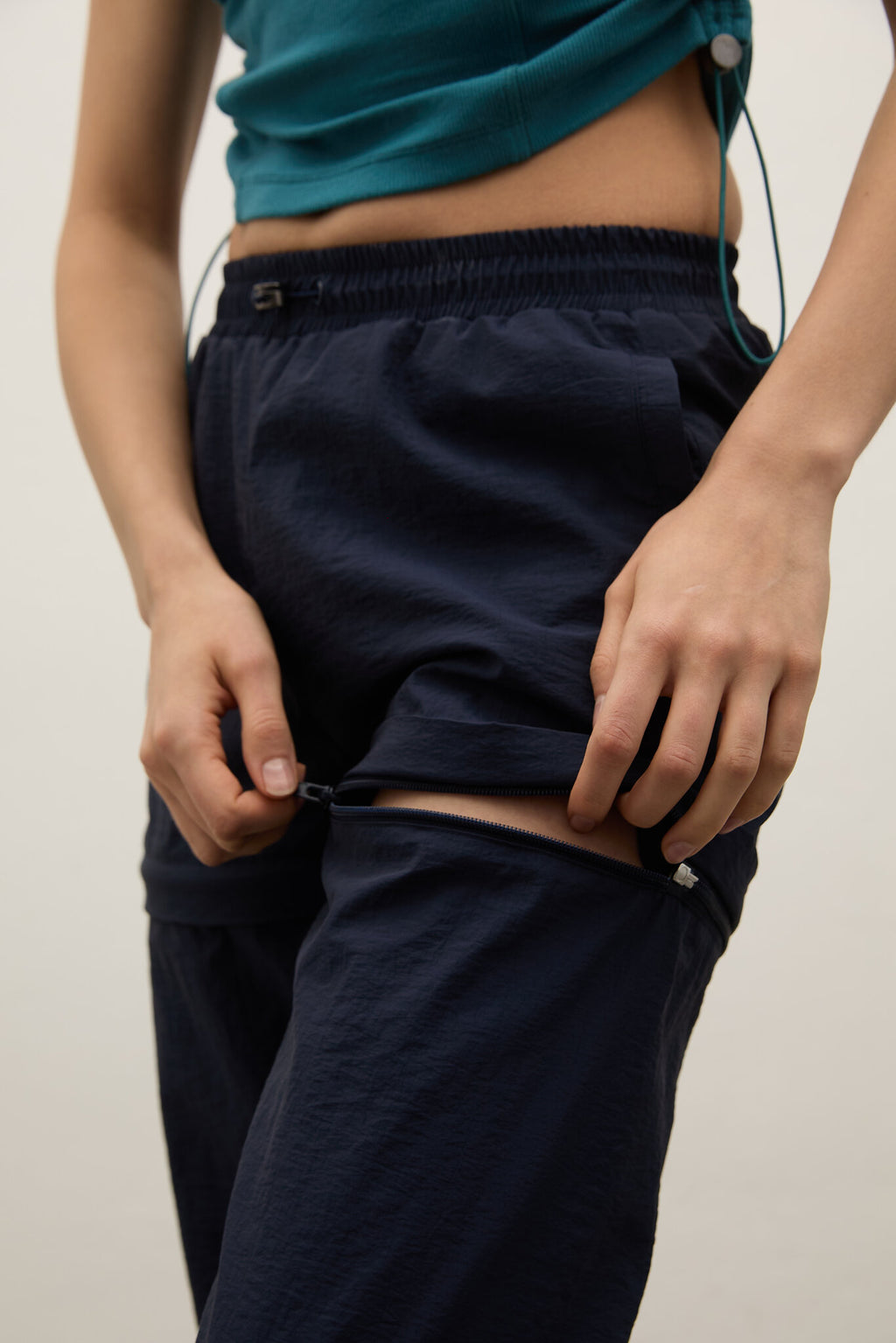 Close-up of a person wearing dark navy pants with a high waist and elastic band, pulling the side seam apart to demonstrate a stretch feature. The person is also wearing a teal cropped top against a plain light background.
