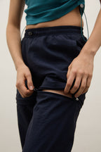 Close-up of a person wearing dark navy pants with a high waist and elastic band, pulling the side seam apart to demonstrate a stretch feature. The person is also wearing a teal cropped top against a plain light background.