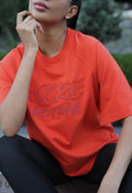 A woman wearing a bright orange short-sleeve t-shirt with 'MORE ACTIVE' embroidered in pink across the chest, paired with black leggings, in an outdoor setting.