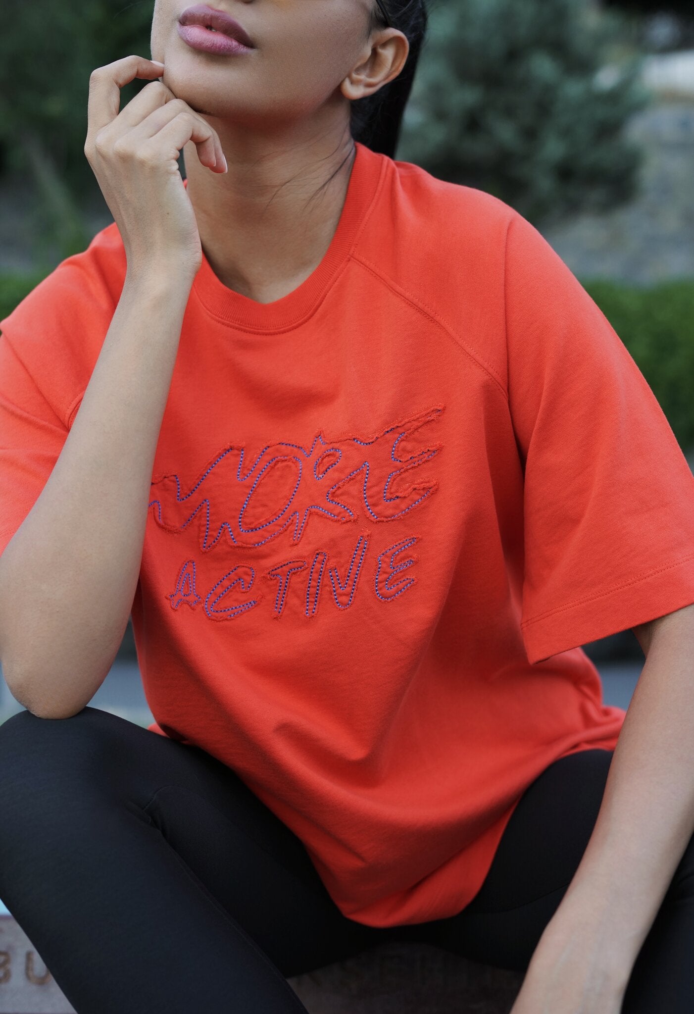 A woman wearing a bright orange short-sleeve t-shirt with 'MORE ACTIVE' embroidered in pink across the chest, paired with black leggings, in an outdoor setting.