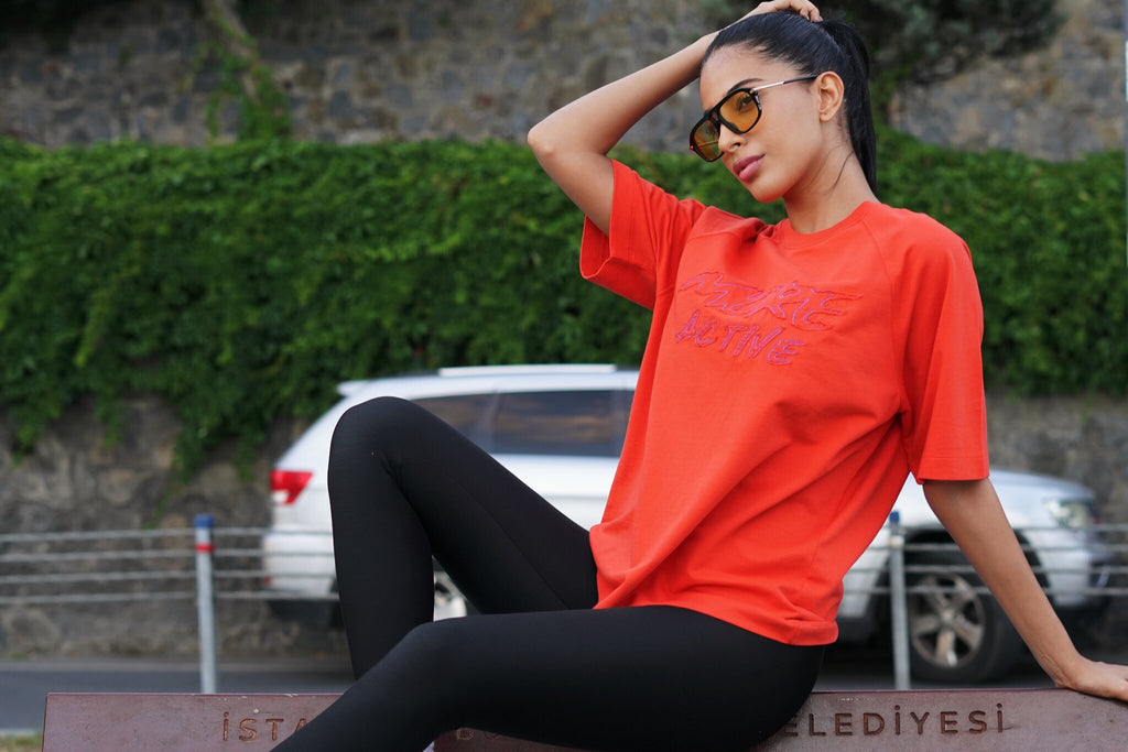 A woman wearing an oversized bright orange-red graphic t-shirt and black leggings sits outdoors against a stone wall with green ivy and a white car in the background.