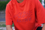 A close-up of a person wearing a bright orange-red raglan t-shirt with 'MORE ACTIVE' embroidered across the chest in a matching red and blue outline stitch.