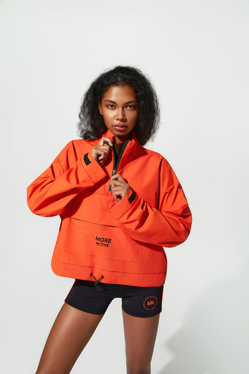 A model wearing a bright orange zip-up jacket with black accents and black athletic shorts, standing against a plain light gray studio background.