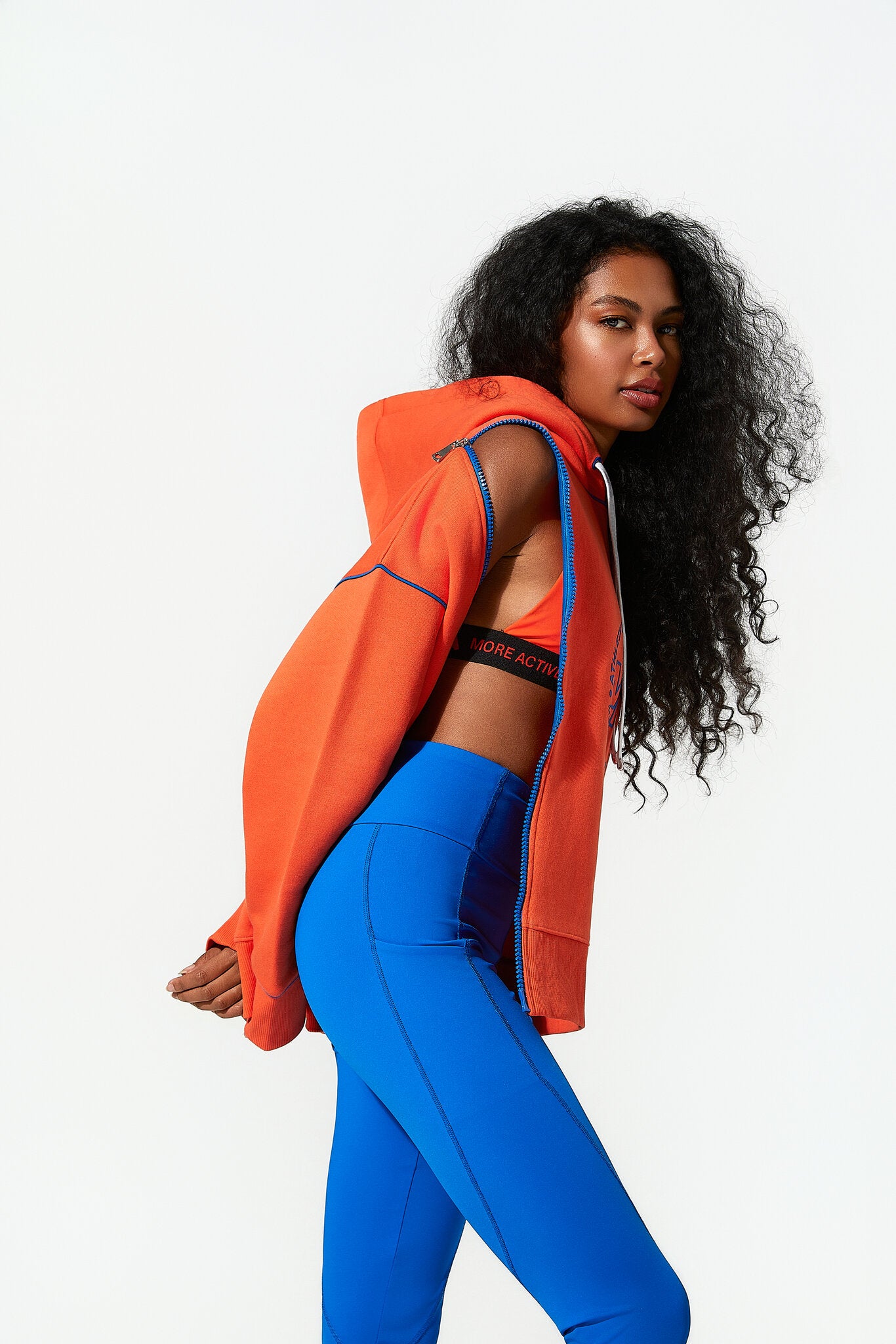 A woman modeling a bright orange zip-up hoodie with blue piping and matching blue leggings against a plain white studio background.