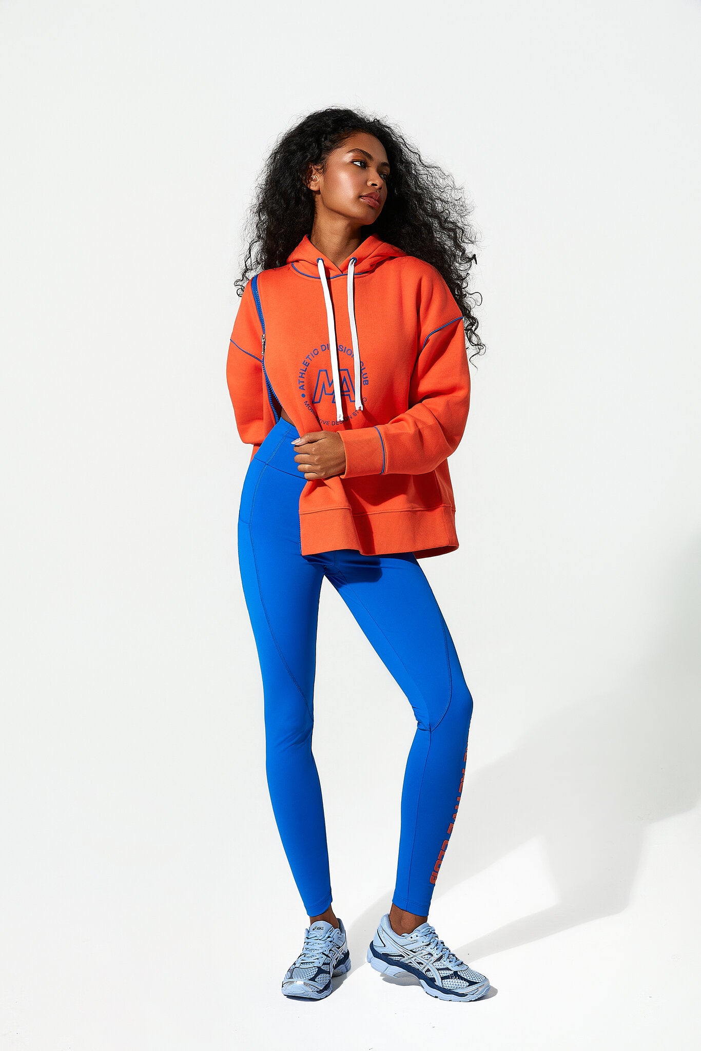 A woman modeling a bright orange cropped hoodie with white drawstrings and a blue graphic print, paired with vibrant blue leggings and light blue running shoes, against a plain white studio background.
