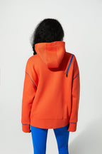 A child viewed from behind wearing a bright orange hooded sweatshirt with blue zipper accents on the shoulders and cuffs, paired with blue leggings, against a plain light gray studio background.
