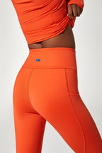 Close-up of a model wearing bright orange high-waisted leggings with a small blue logo on the waistband, paired with a matching orange top against a light gray studio background.