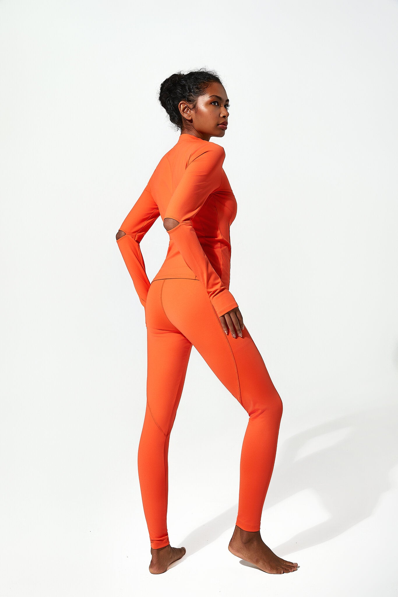 A woman modeling a bright orange two-piece athletic set consisting of a long-sleeve mock-neck top and matching leggings, shown from a side-back angle against a clean white studio background.