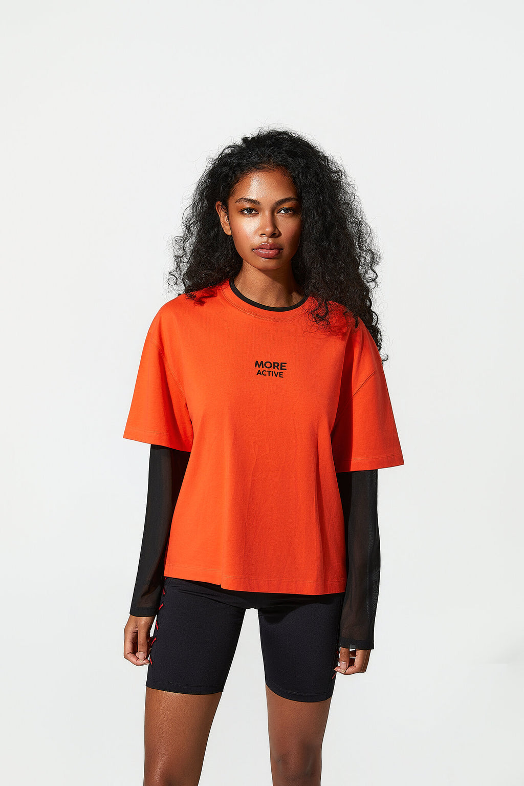 A woman modeling an oversized bright orange short-sleeve t-shirt with black trim and the words 'MORE ACTIVE' printed on the chest, layered over a black long-sleeve shirt and black bike shorts, against a plain white studio background.