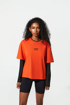 A woman modeling an oversized bright orange short-sleeve t-shirt with black trim and the words 'MORE ACTIVE' printed on the chest, layered over a black long-sleeve shirt and black bike shorts, against a plain white studio background.