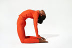 A model in a bright orange athletic tracksuit performing a deep backbend yoga pose against a clean white background.