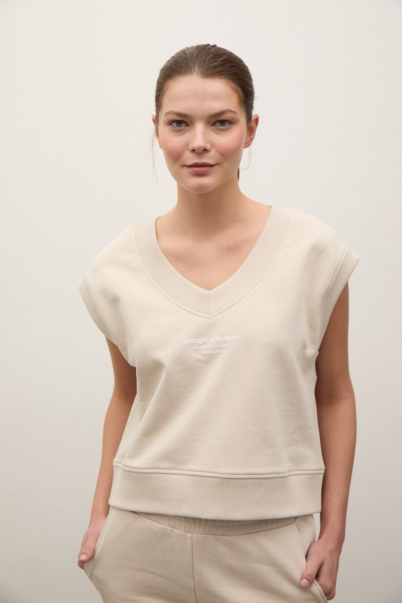 A woman modeling a cream-colored sleeveless V-neck sweatshirt with a small tonal embroidered logo on the chest, paired with matching cream sweatpants against a plain off-white background.