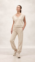 A woman modeling a matching beige loungewear set consisting of a sleeveless V-neck sweatshirt and jogger pants, paired with beige sneakers, against a neutral studio backdrop.