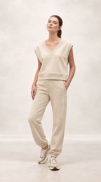 A woman modeling a matching beige loungewear set consisting of a sleeveless V-neck sweatshirt and jogger pants, paired with beige sneakers, against a neutral studio backdrop.