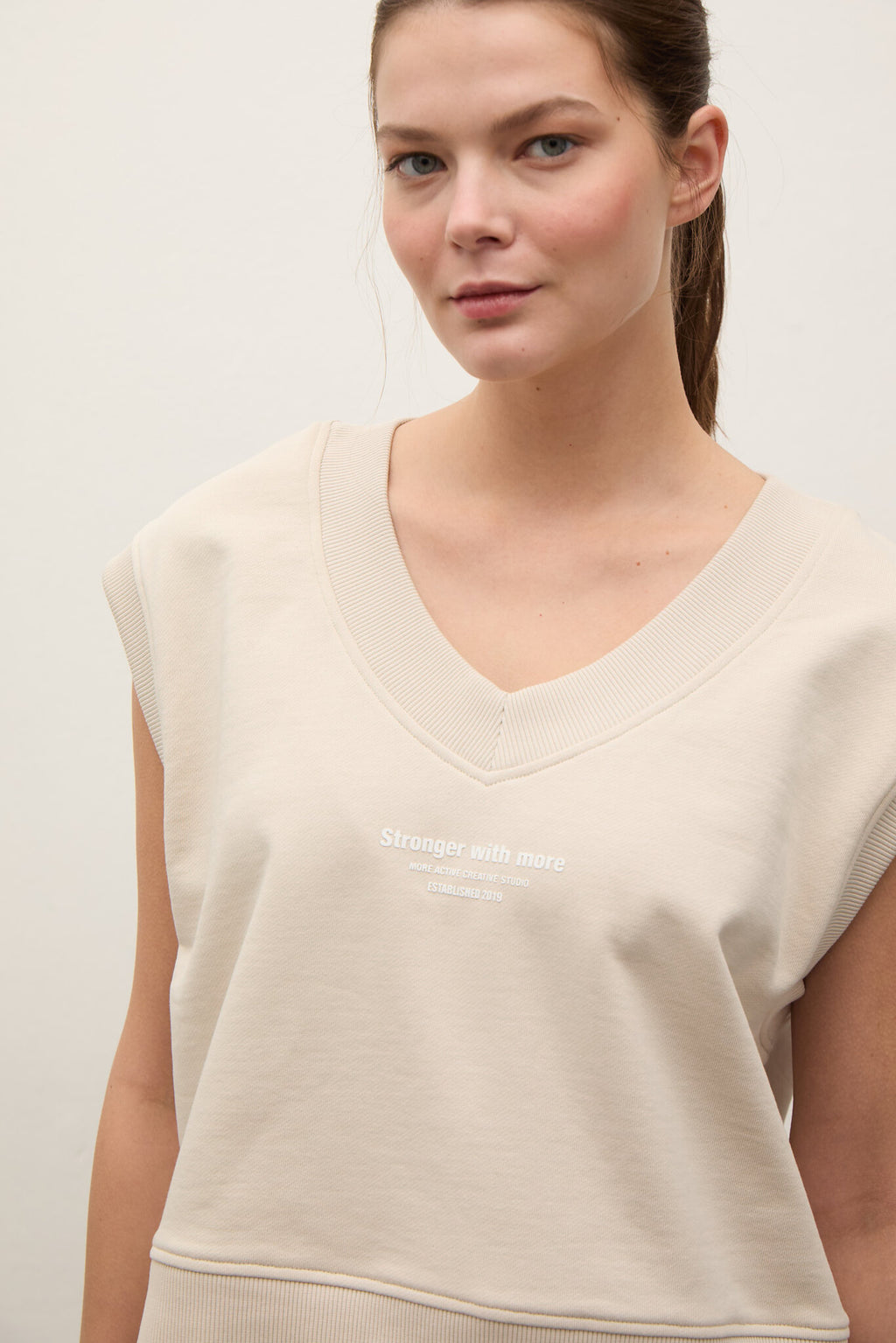 A woman wearing a light beige cropped V-neck sweatshirt with short sleeves and white text on the chest that reads 'Stronger with more'.