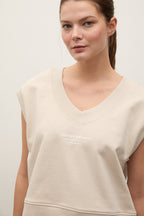 A woman wearing a light beige cropped V-neck sweatshirt with short sleeves and white text on the chest that reads 'Stronger with more'.