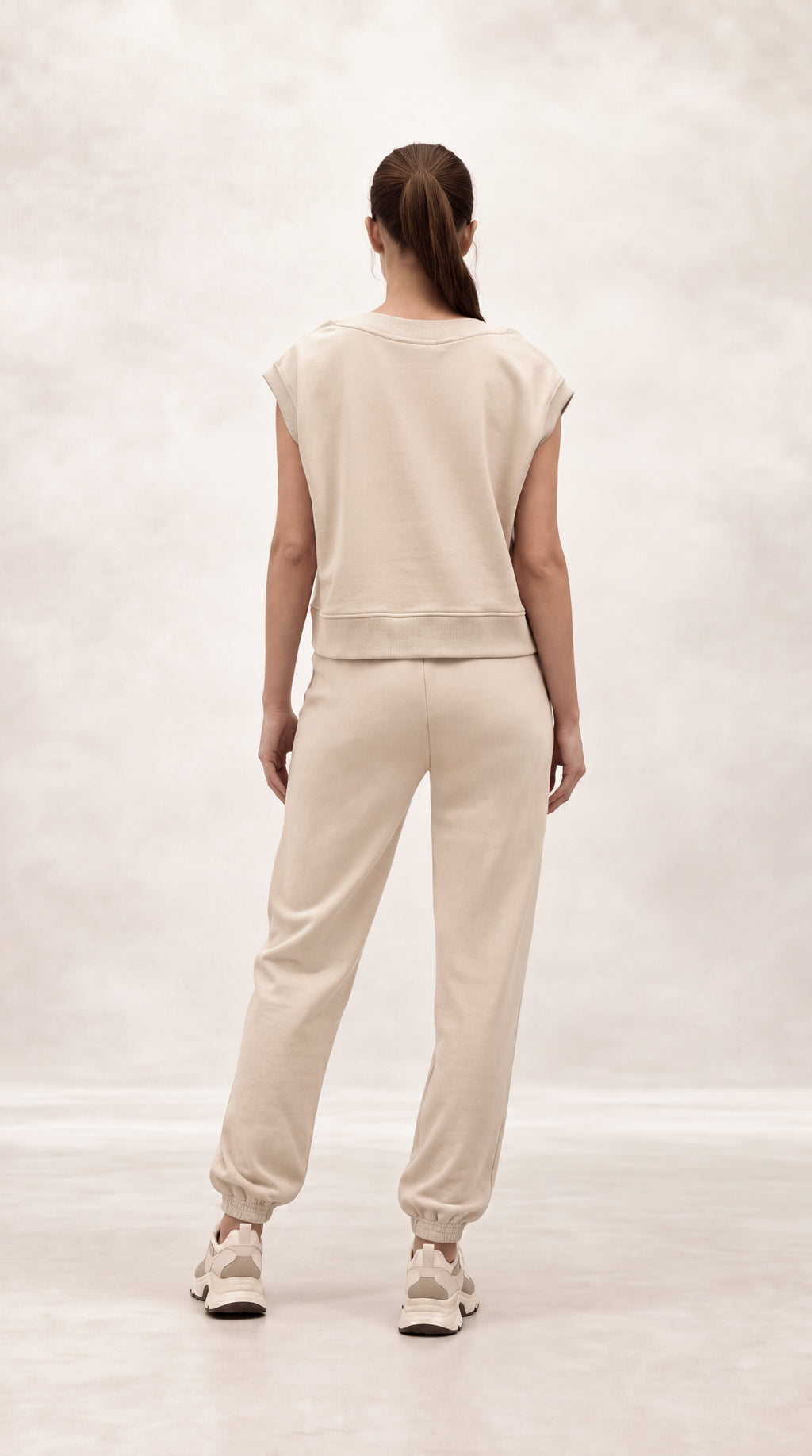 A woman viewed from behind wearing a matching beige cropped sweatshirt and jogger pants set, paired with light-colored sneakers, against a soft, light gray studio backdrop.