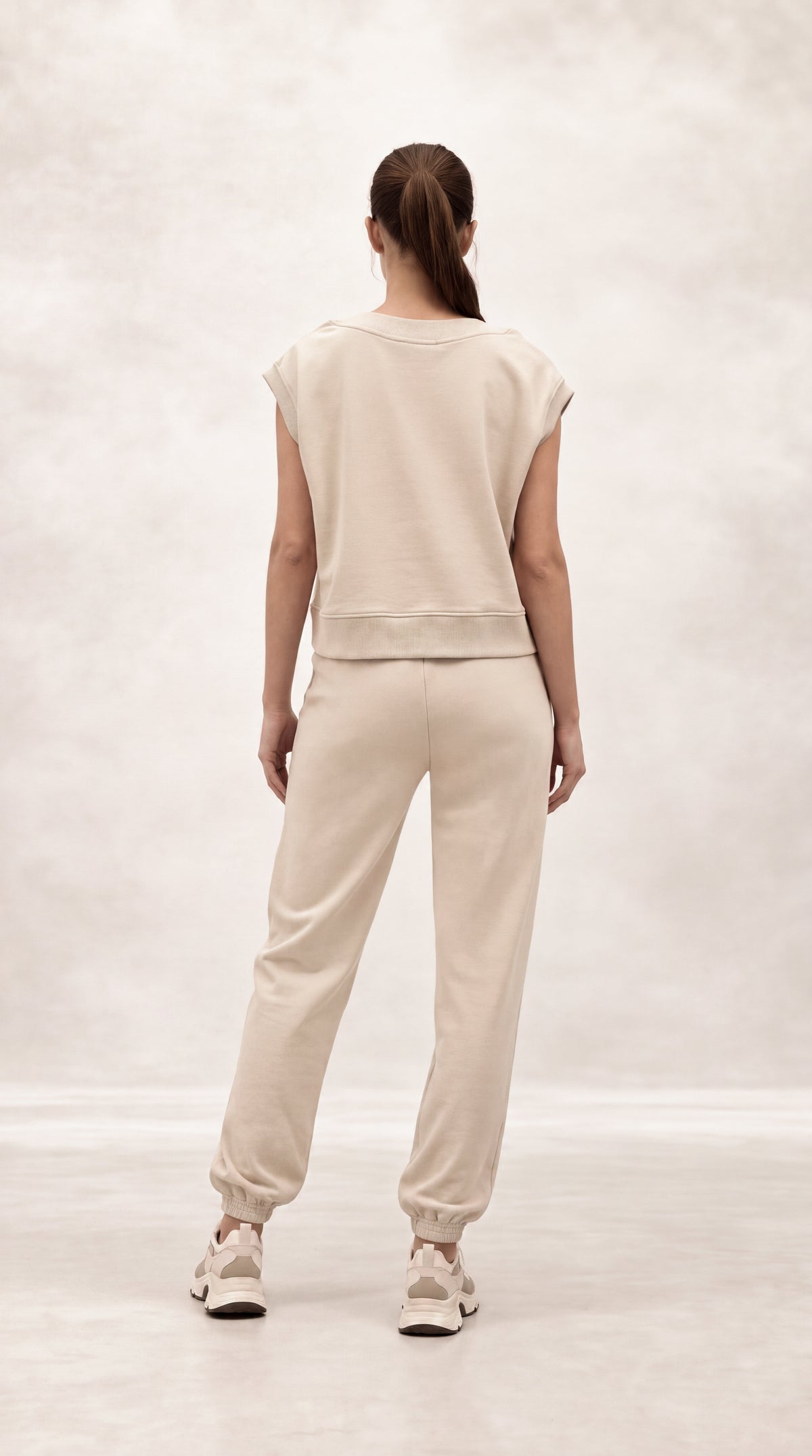 A woman viewed from behind wearing a matching beige cropped sweatshirt and jogger pants set, paired with light-colored sneakers, against a soft, light gray studio backdrop.