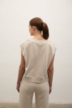 A woman with her hair in a ponytail is shown from behind, wearing a light beige sleeveless top and matching beige pants against a plain off-white wall.