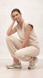 A woman crouching in a studio setting, wearing a matching beige sleeveless top and jogger pants set with beige sneakers.