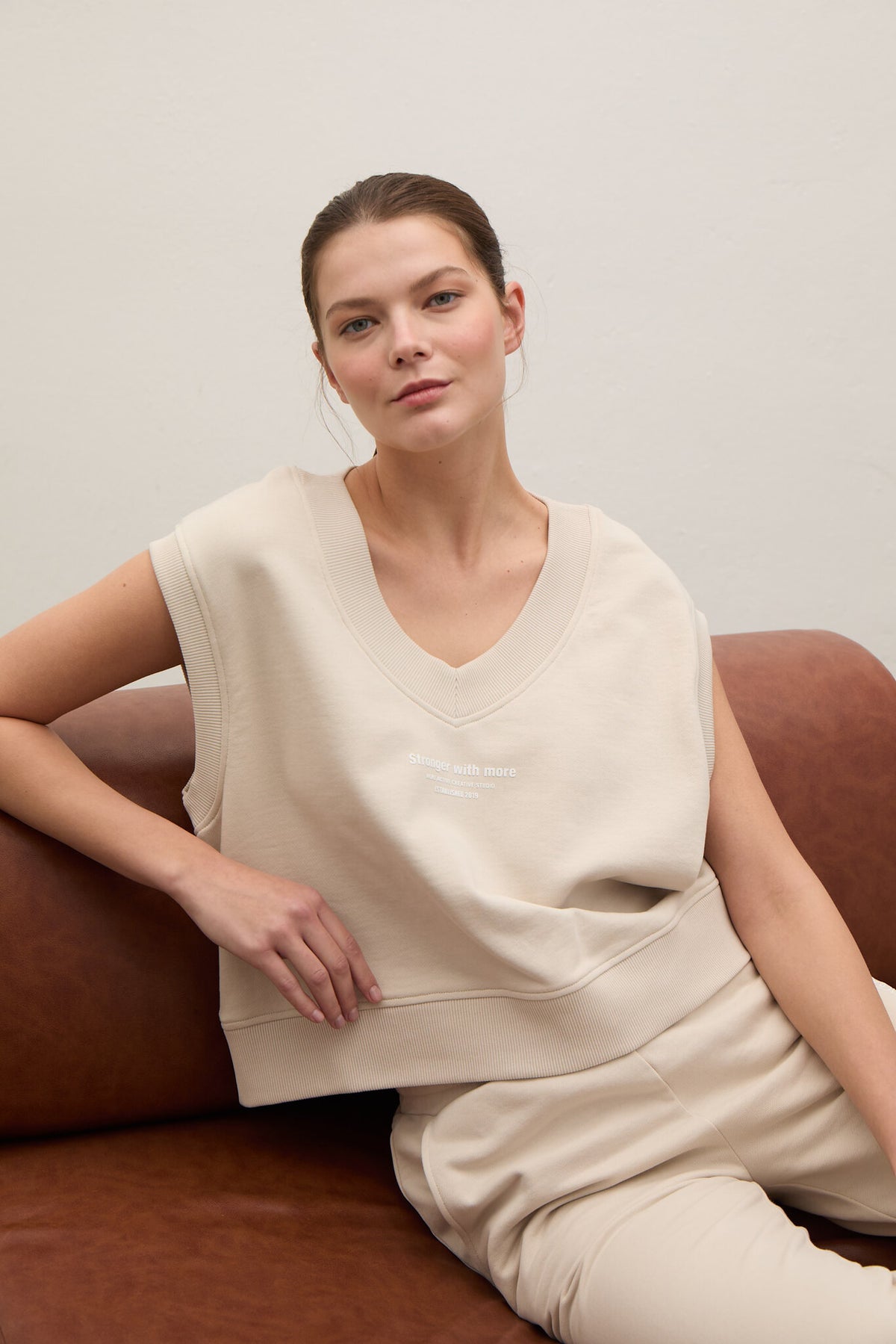 A woman wearing a beige sleeveless V-neck sweatshirt and matching beige pants, seated on a brown leather couch against a light gray wall.