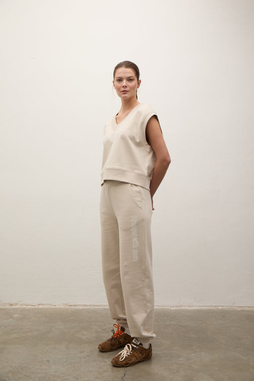 A model stands in a minimalist studio setting wearing a beige sleeveless V-neck sweatshirt and matching beige sweatpants with vertical text on the side, paired with brown sneakers.