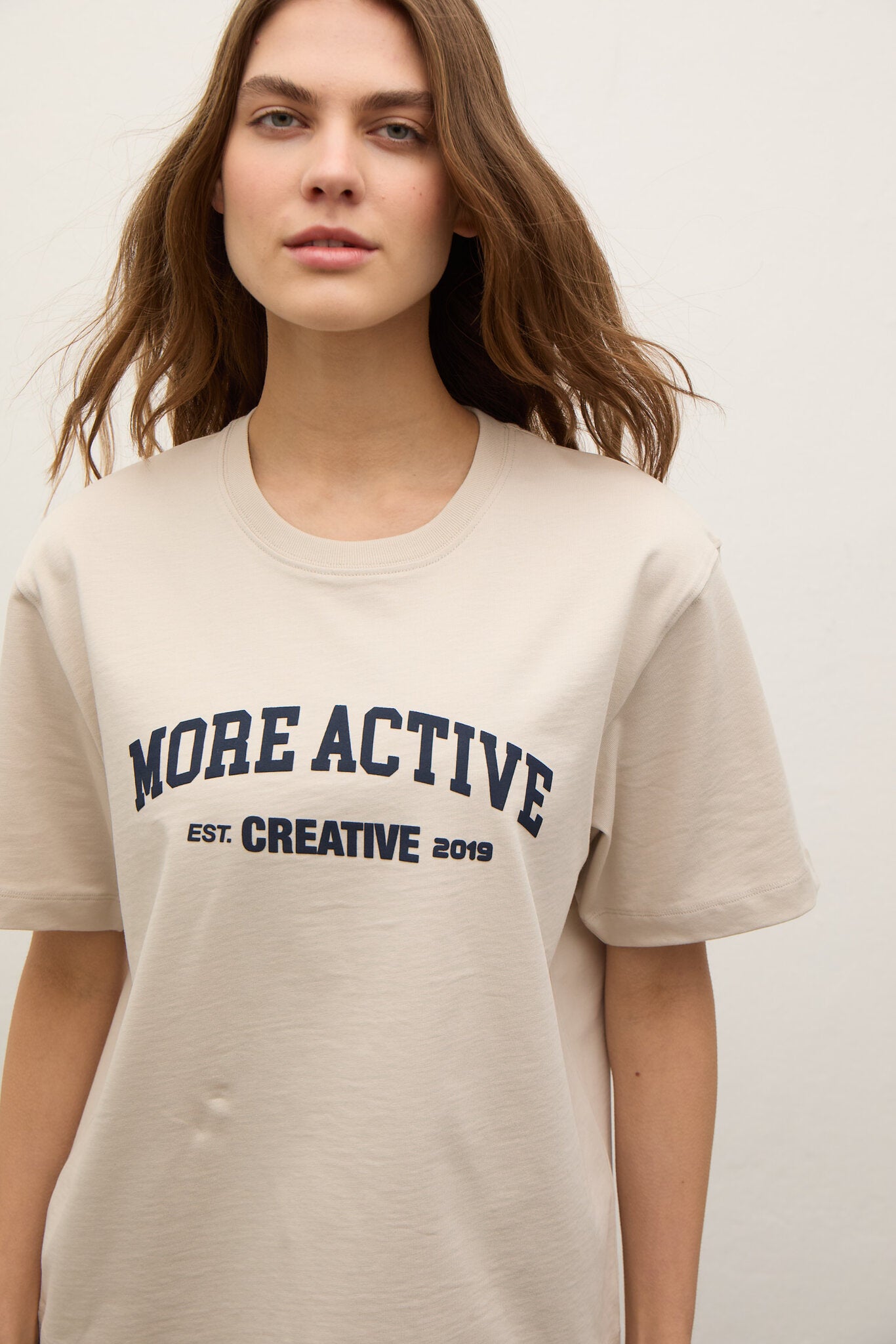 A woman wearing a light beige crewneck t-shirt with navy blue text that reads 'MORE ACTIVE EST. CREATIVE 2019' on the front, set against a plain light background.