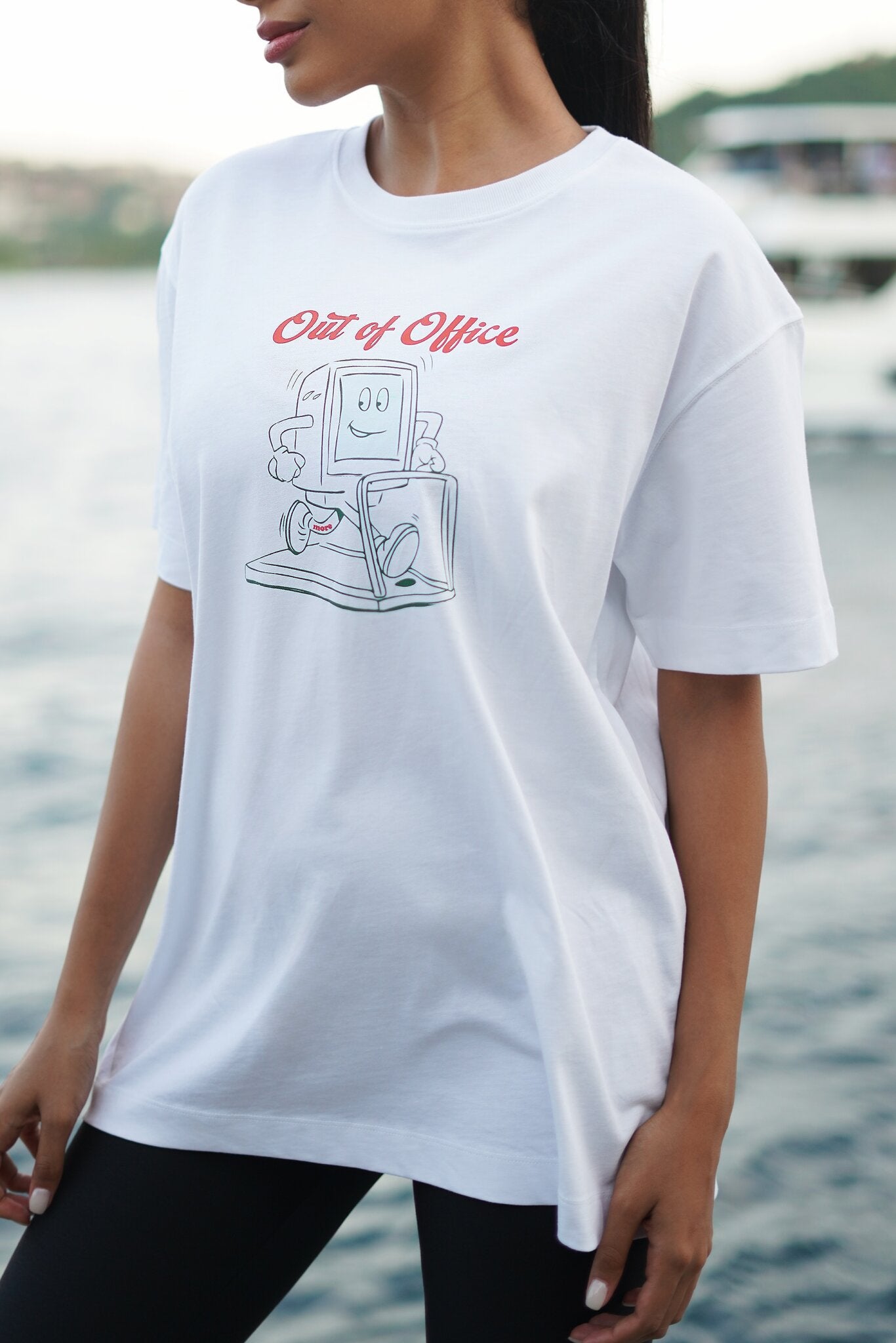 A woman wearing a white oversized graphic t-shirt with a retro computer illustration and the text 'Out of Office' in red, set against a blurred background of water and a boat.