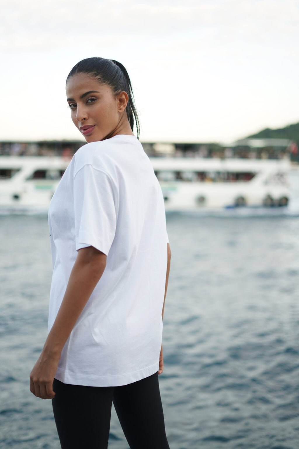 A woman wearing an oversized white t-shirt and black pants, photographed from behind in an outdoor setting with water and boats in the background.