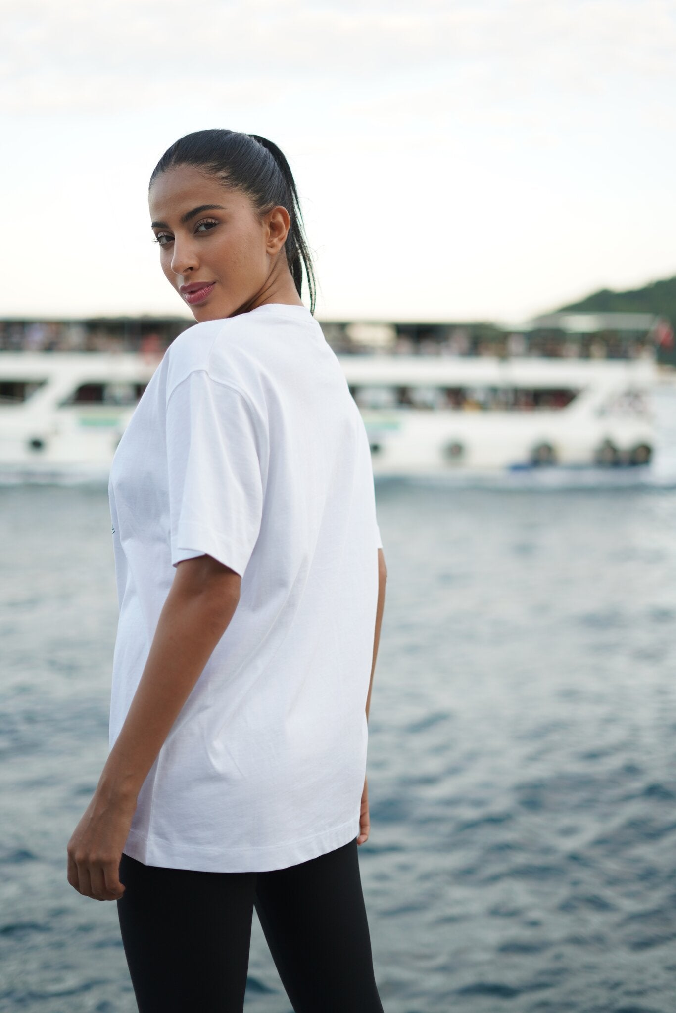 A woman wearing an oversized white t-shirt and black pants, photographed from behind in an outdoor setting with water and boats in the background.