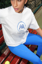 A woman sitting on a red wooden bench wearing a white athletic t-shirt with a blue circular logo that reads 'ATHLETIC DIVISION CLUB' and 'MA', paired with bright blue leggings.
