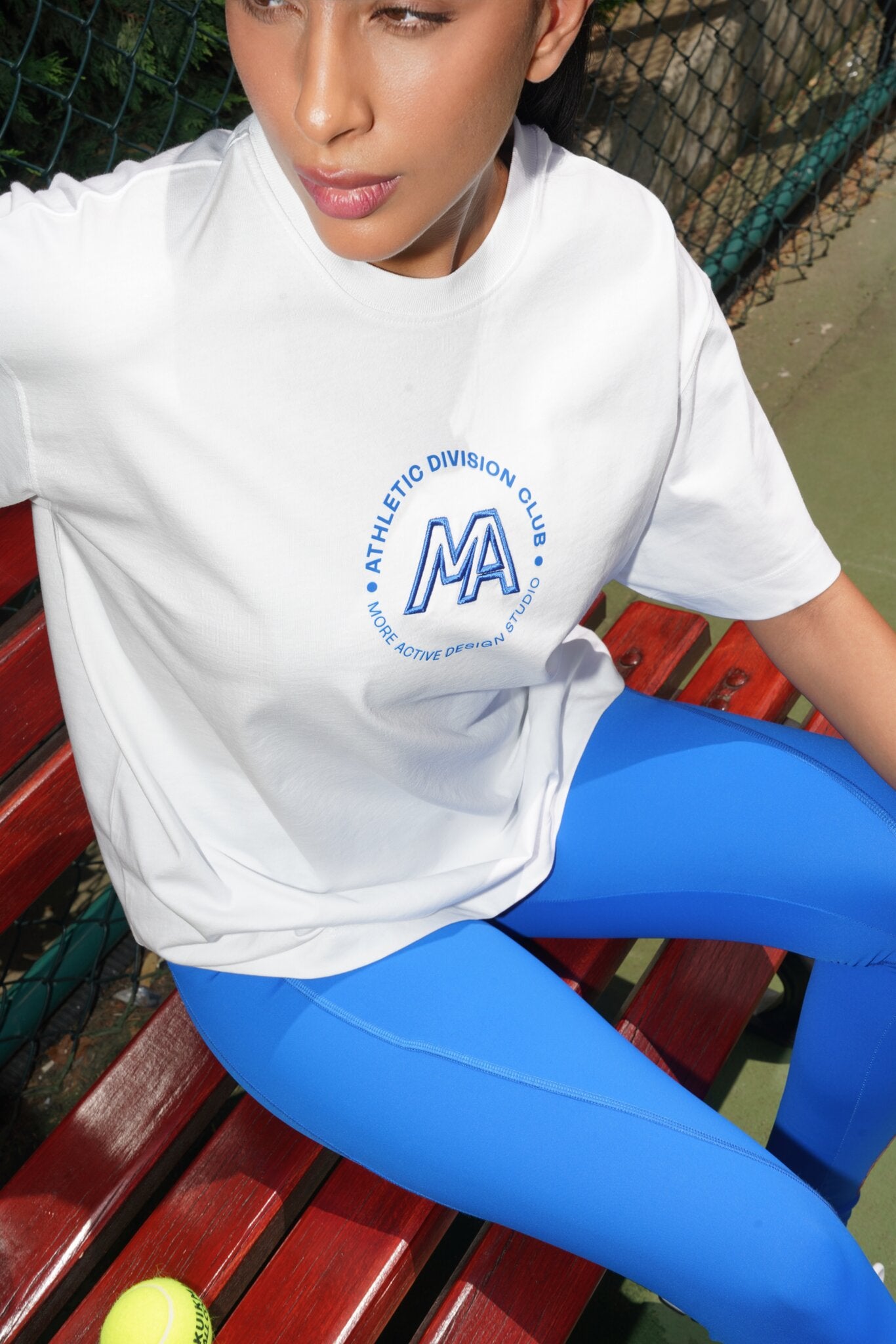 A woman sitting on a red wooden bench wearing a white athletic t-shirt with a blue circular logo that reads 'ATHLETIC DIVISION CLUB' and 'MA', paired with bright blue leggings.