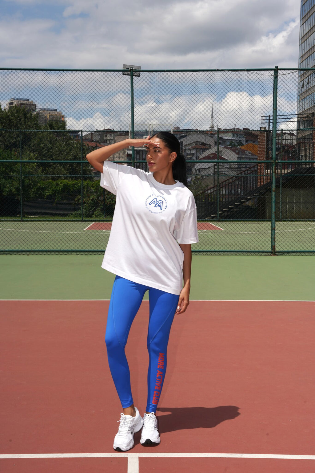 A woman stands on an outdoor tennis court wearing a white graphic t-shirt, bright blue leggings with red text, and white sneakers. The background shows a green chain-link fence, other court surfaces, and a cloudy sky.