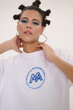 A woman wearing a white crewneck t-shirt with a blue circular graphic on the chest that reads 'ATHLETICO DIVISION CLUB' and 'MA MORE ACTIVE DESIGN STUDIO'. She is posing against a clean, off-white background.