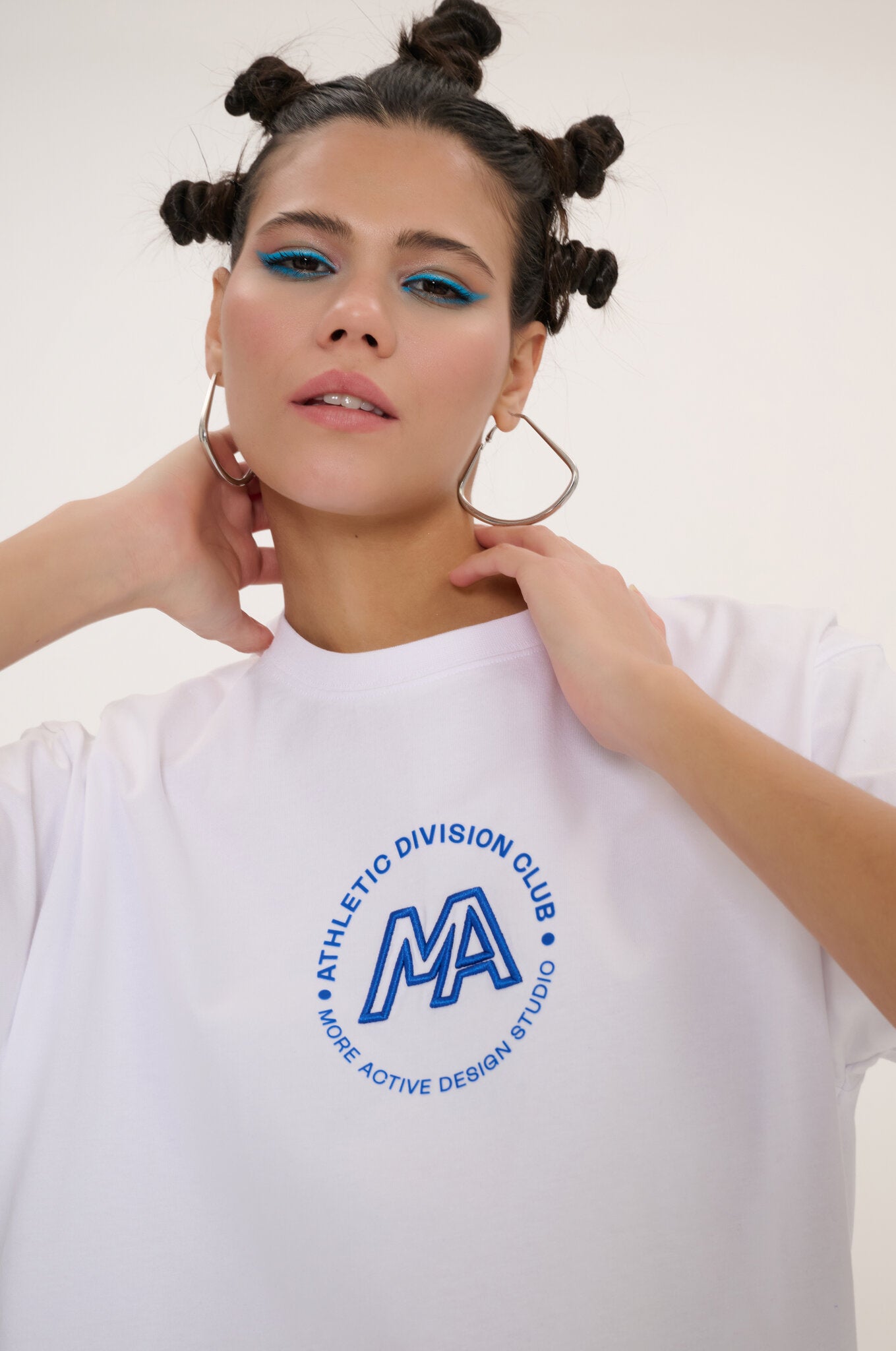 A woman wearing a white crewneck t-shirt with a blue circular graphic on the chest that reads 'ATHLETICO DIVISION CLUB' and 'MA MORE ACTIVE DESIGN STUDIO'. She is posing against a clean, off-white background.