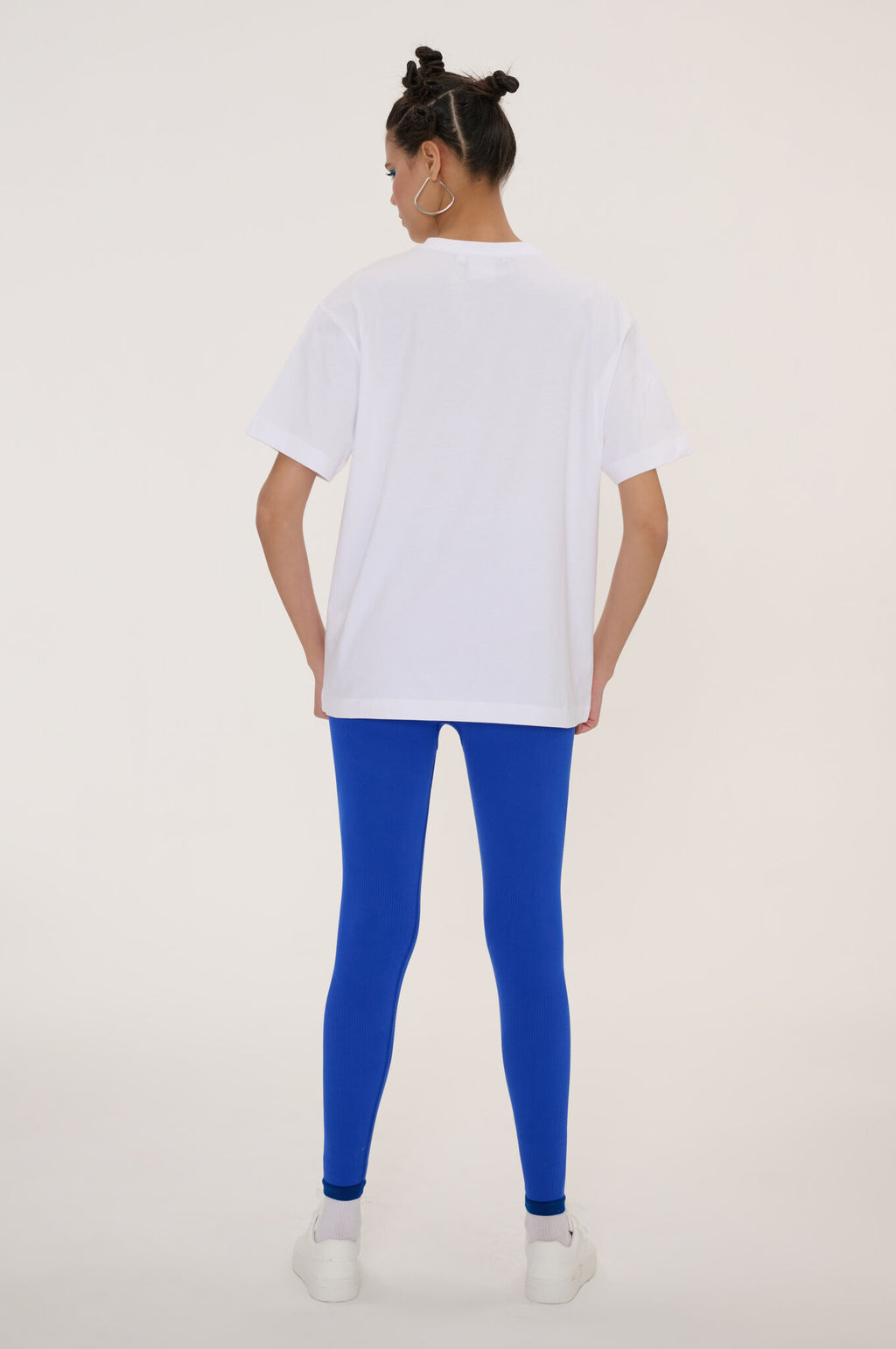A woman shown from behind wearing a white oversized short-sleeve t-shirt, bright royal blue leggings, and white sneakers against a plain light beige studio background.