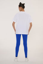 A woman shown from behind wearing a white oversized short-sleeve t-shirt, bright royal blue leggings, and white sneakers against a plain light beige studio background.