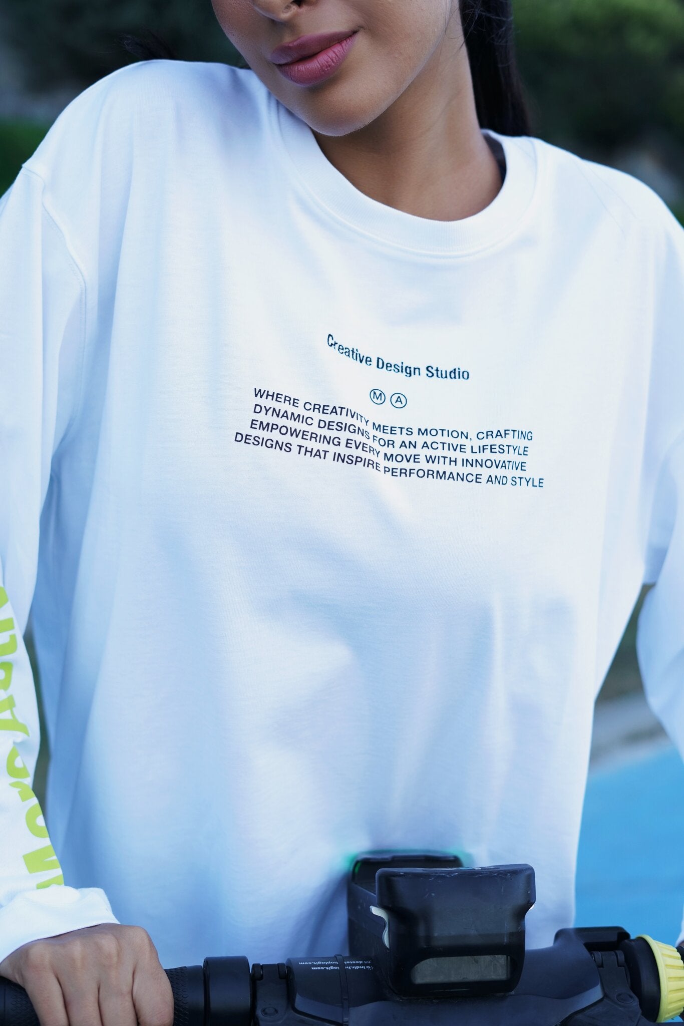 A close-up of a person wearing a white long-sleeve t-shirt with black printed text on the chest, holding the handlebars of a bicycle outdoors.