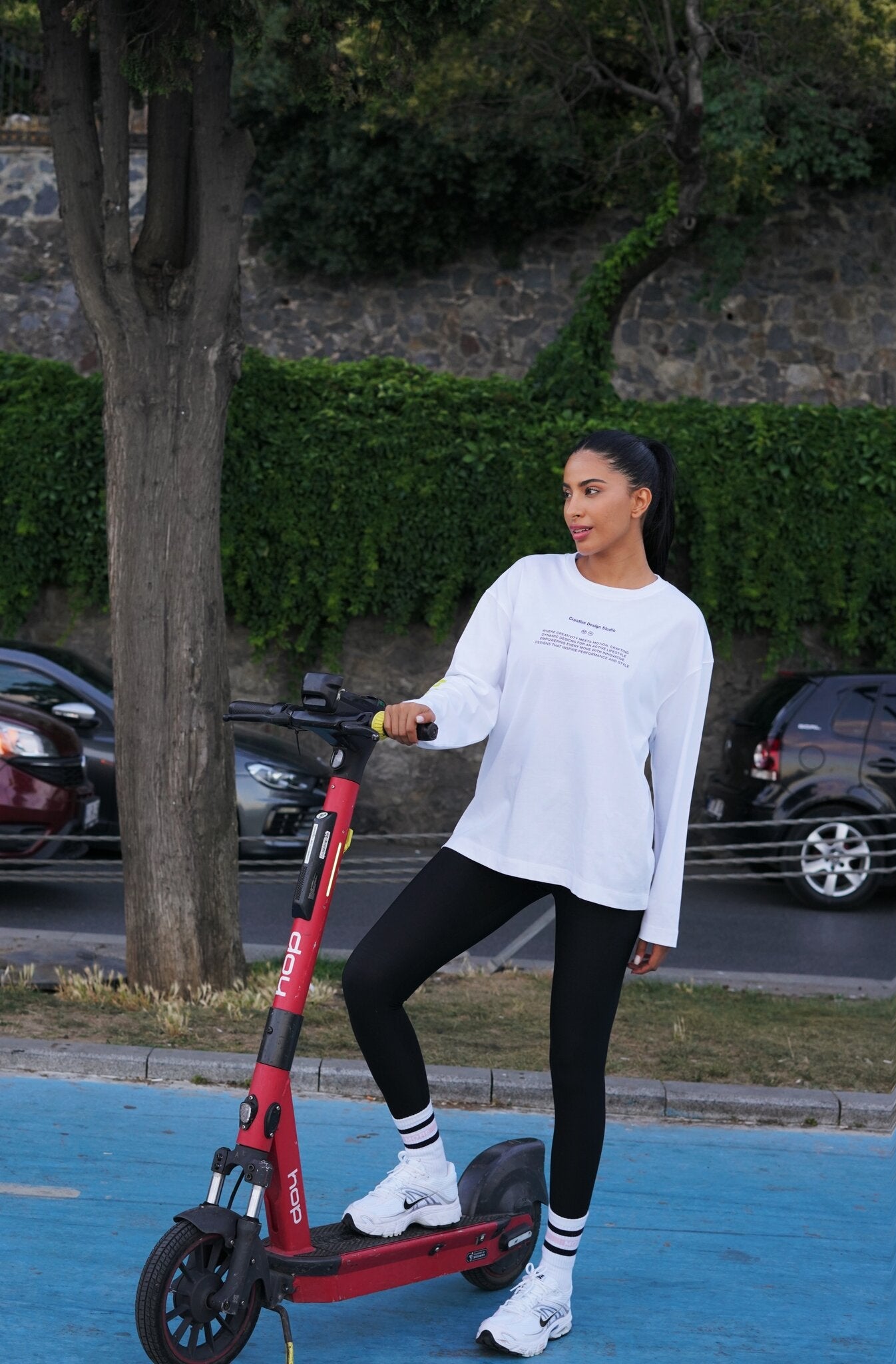 A woman stands on a red electric scooter on a blue paved path outdoors, wearing a white long-sleeve t-shirt, black leggings, and white sneakers. Trees, parked cars, and a stone wall are visible in the background.
